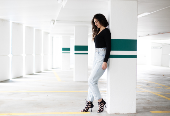 Frau in schwarzem Top und Jeans steht an einer Wand in einem hellen, leeren Parkhaus.