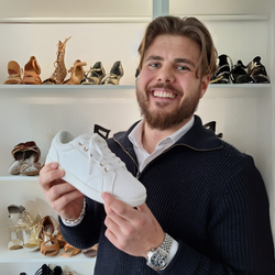 Lächelnder Mann mit weißem Turnschuh in der Hand in einem Schuhgeschäft, im Hintergrund Regale mit verschiedenen Schuhen.