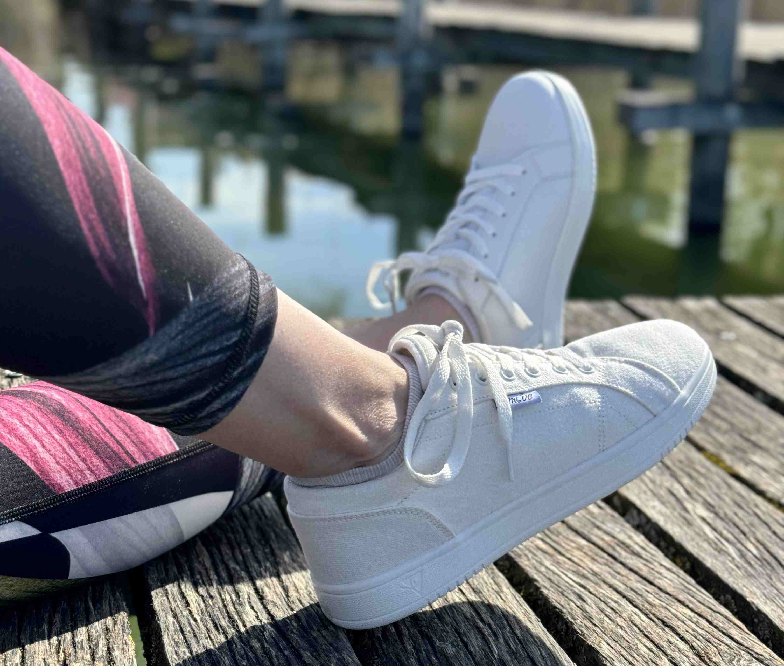 Person in weißen Turnschuhen und bunten Leggings, die auf einem Holzsteg in der Nähe von Wasser sitzt.